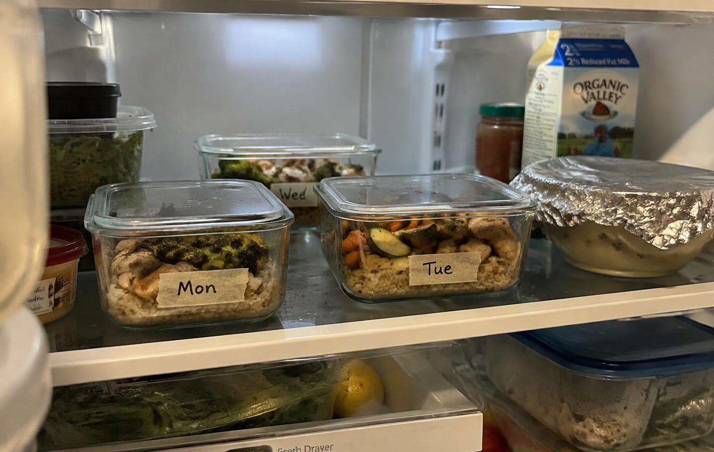 Labelled glass meal-prep containers of chicken, rice and vegetables stacked on a refrigerator shelf